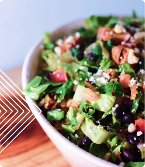 A bowl of salad with lettuce, tomatoes, black beans, cheese, and croutons, placed on a wooden surface with a geometric design on the left.