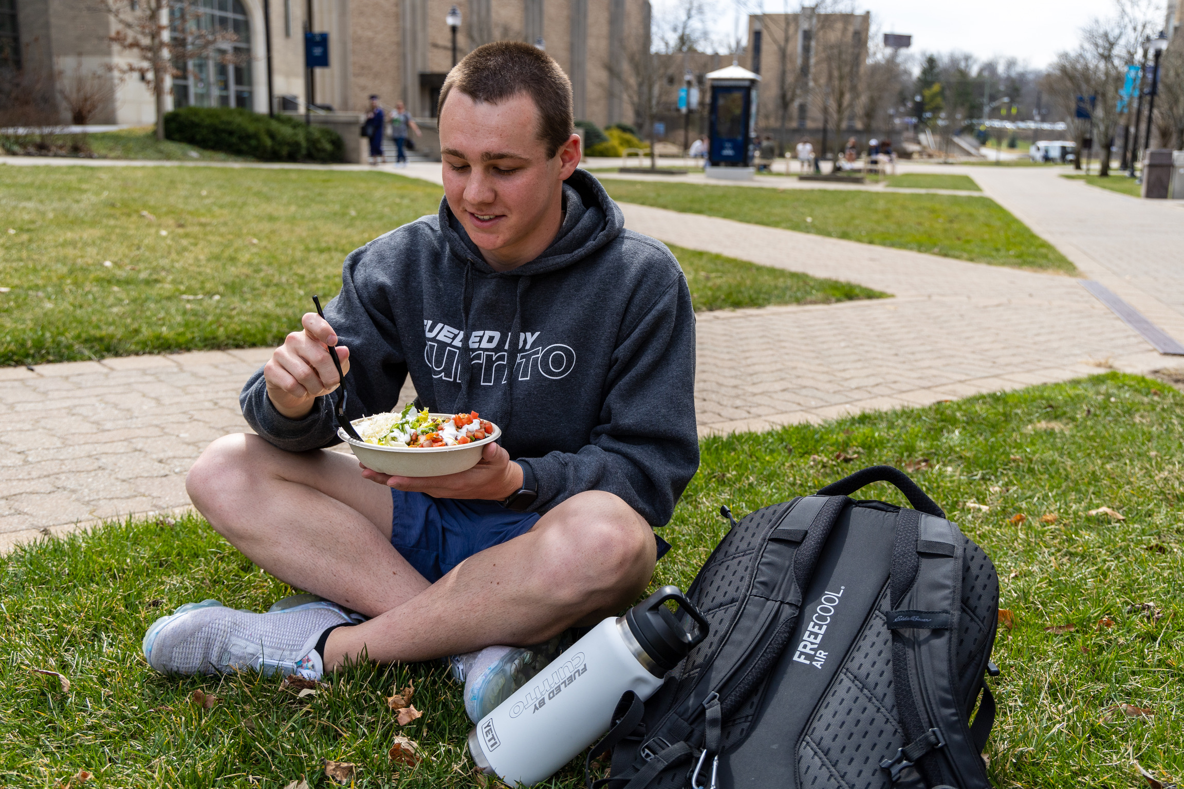 A man sitting on the ground of a school campus eating a currito bowl with his backpack next to him.